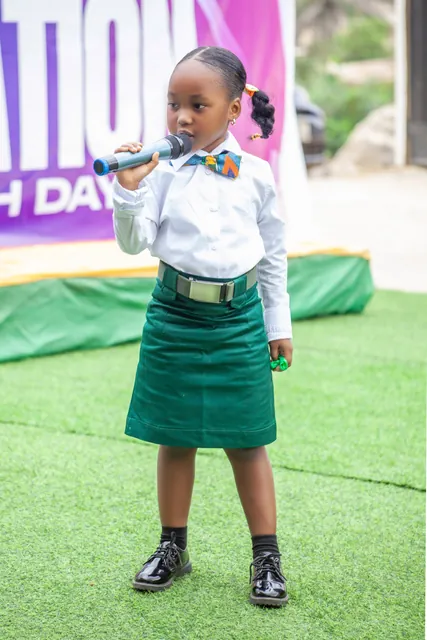 A young pupil in the green and white school uniform speaking into a microphone at an outdoor school event