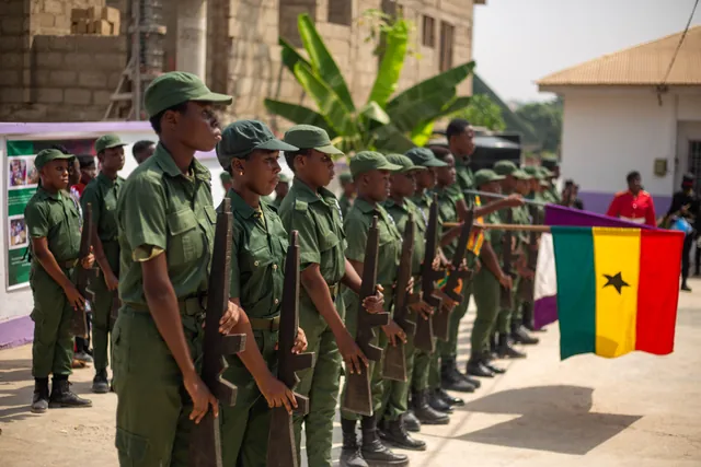 Pupils in green parade uniform standing at attention on the school grounds beside the flag of Ghana